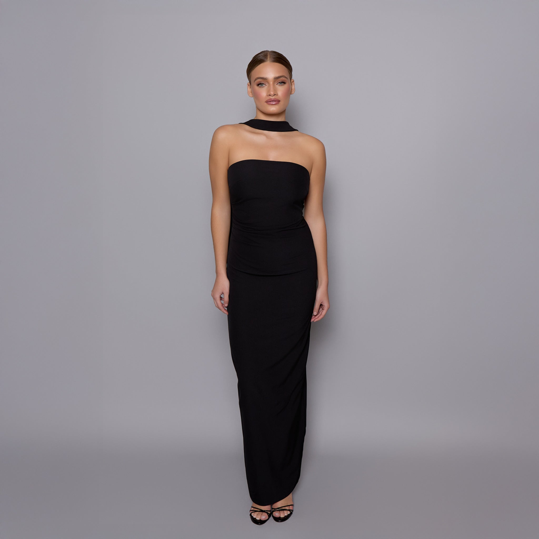 Image of a model wearing the vesper tiffany maxi dress in black featuring a neck scarf and ruched waist on a grey background