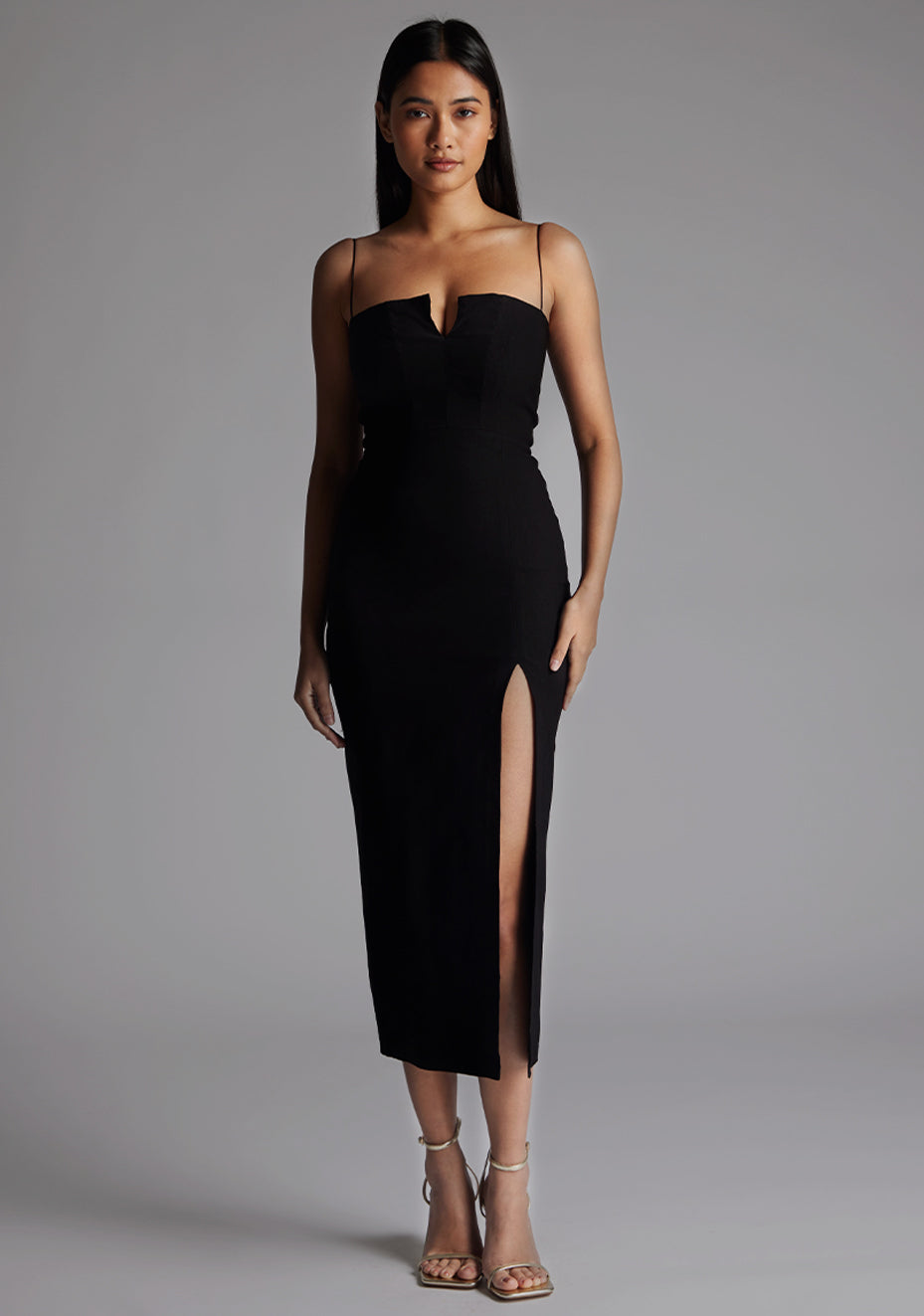 Model wearing a black midaxi dress with thin straps, a structured sweetheart-style neckline, and a thigh-high front split, standing against a grey studio background.