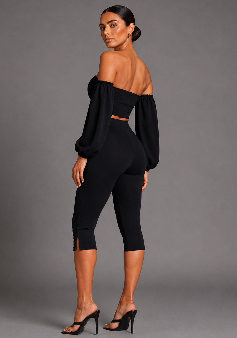 Side back image of a model wearing the VSPR Belle Sweetheart Neckline Puff Sleeve Black Top highlighting the structured puff sleeves, smooth stretch fabric, and flattering body-hugging shape, The design paired with Capri Black Cropped Trousers, standing against a grey studio backdrop.