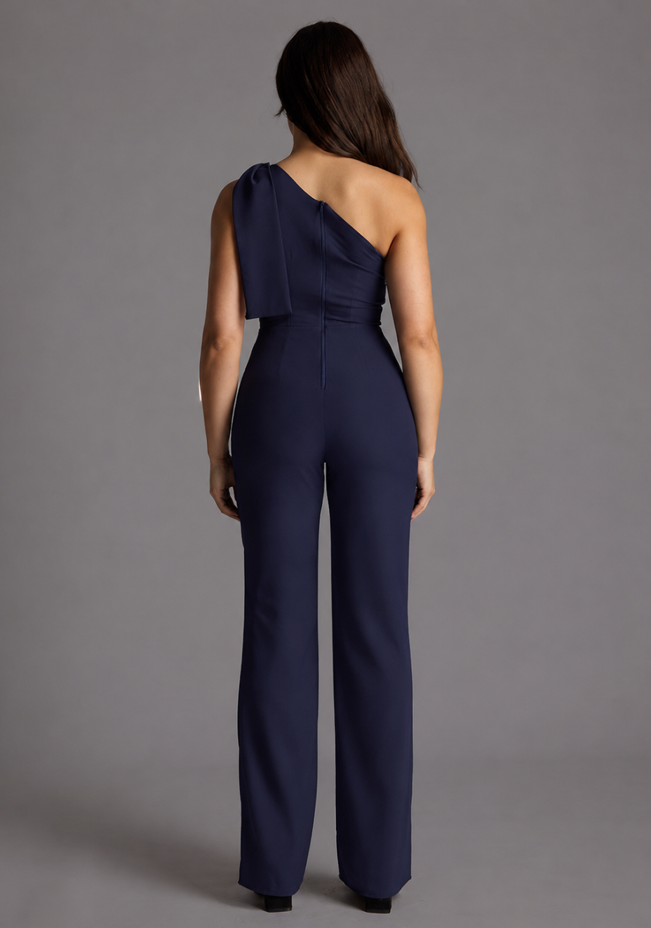 Back image of a model wearing the VSPR Gracie Petite One Shoulder Wide Leg Jumpsuit in Navy, highlighting the clean tailored back and elegant wide-leg silhouette against a grey studio backdrop.