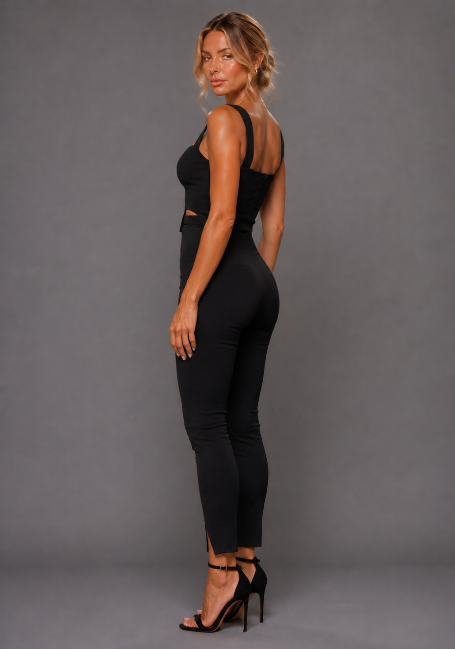 Side back image of a model wearing the VSPR Juno Sweetheart Black Crop Top with wide supportive straps and a sculpted fit, paired with the VSPR Raven Slim Leg High Waisted Black Trousers highlighting the clean tailored fit and streamlined silhouette, against a grey studio backdrop.