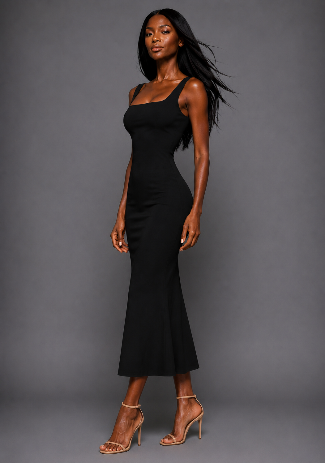 Side front image of a model wearing the VSPR Marianna Square Neckline Fishtail Black Midaxi Dress highlighting the contoured bodice, figure-enhancing fit, and softly flared fishtail skirt against a grey studio backdrop.