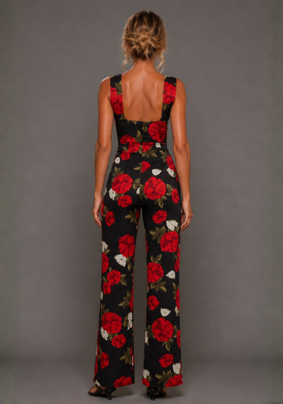 Back image of a model wearing the VSPR Noelle V Neckline Black Floral Wide Leg Jumpsuit featuring a low scoop back, tailored waist, and full-length wide leg silhouette against a grey studio backdrop.