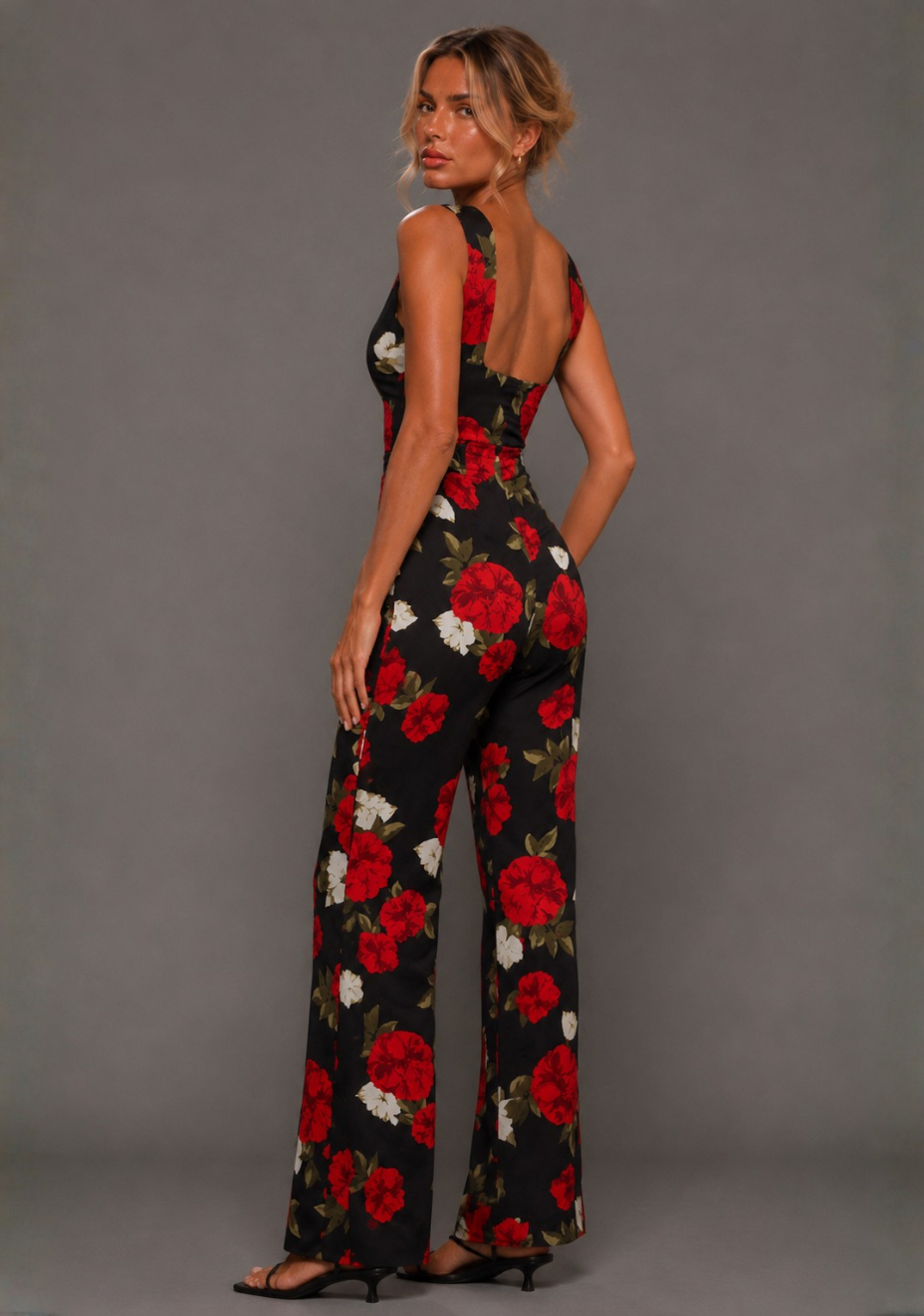 Side back image of a model wearing the VSPR Noelle V Neckline Black Floral Wide Leg Jumpsuit showcasing the low scoop back, flowing wide leg silhouette, and bold floral print against a grey studio backdrop.