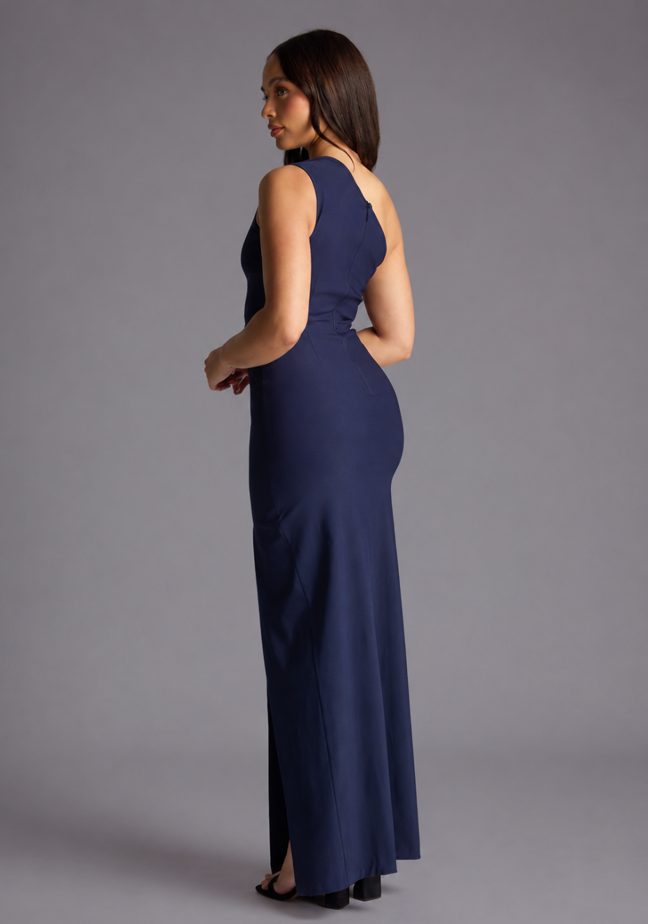 Woman wearing the Vesper Petite Wren navy one shoulder cut out maxi dress against a grey background, side back view