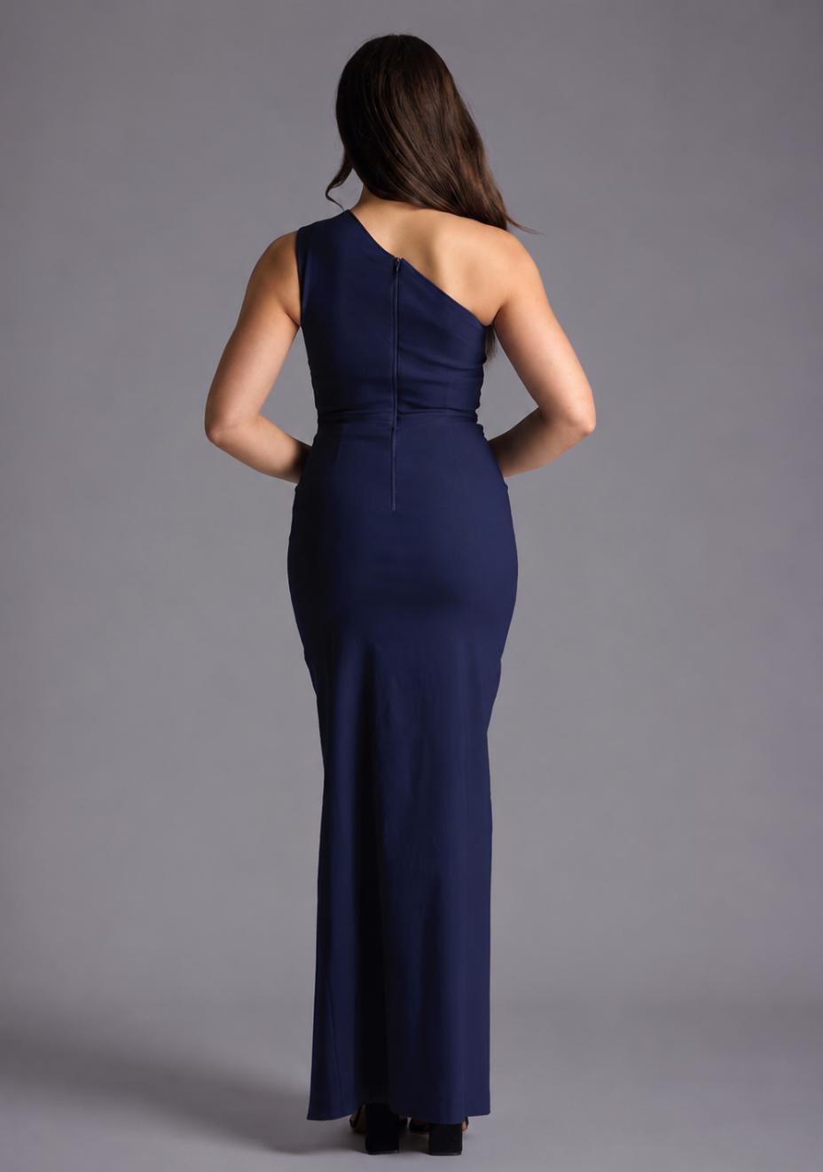 Woman wearing the Vesper Petite Wren navy one shoulder cut out maxi dress against a grey background, back view