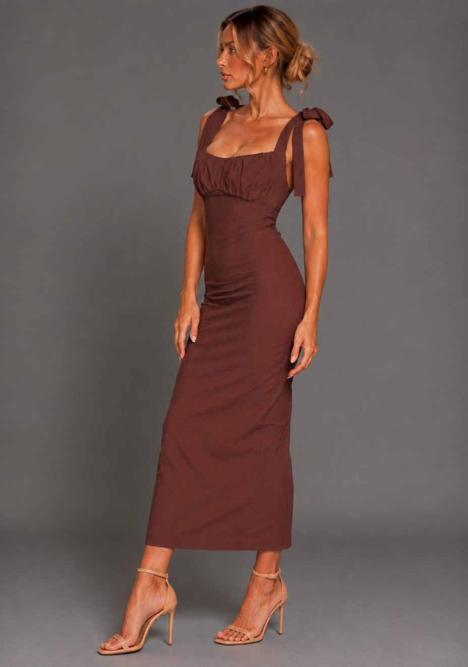 Side front image of a model wearing the VSPR Rapunzel Ruched Bust Bodycon Chocolate Midaxi Dress in Chocolate, highlighting its body-skimming silhouette, ruched bust detail, delicate straps, and flattering midaxi length against a grey studio backdrop.