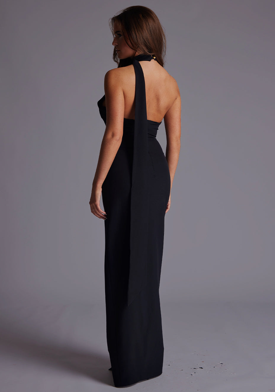Side back image of a model wearing the Vesper Ariella High Neck Maxi Dress in Black, revealing the chic open back and elegant halter neckline with long draped tie detail.