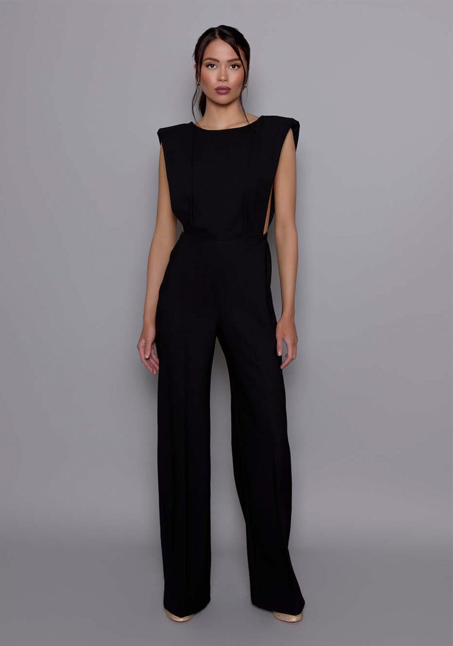 Model wearing the VSPR Bronte Black open-back Jumpsuit with a sharp shoulder line, structured bodice, and wide-leg trousers, standing against a neutral gray studio backdrop.