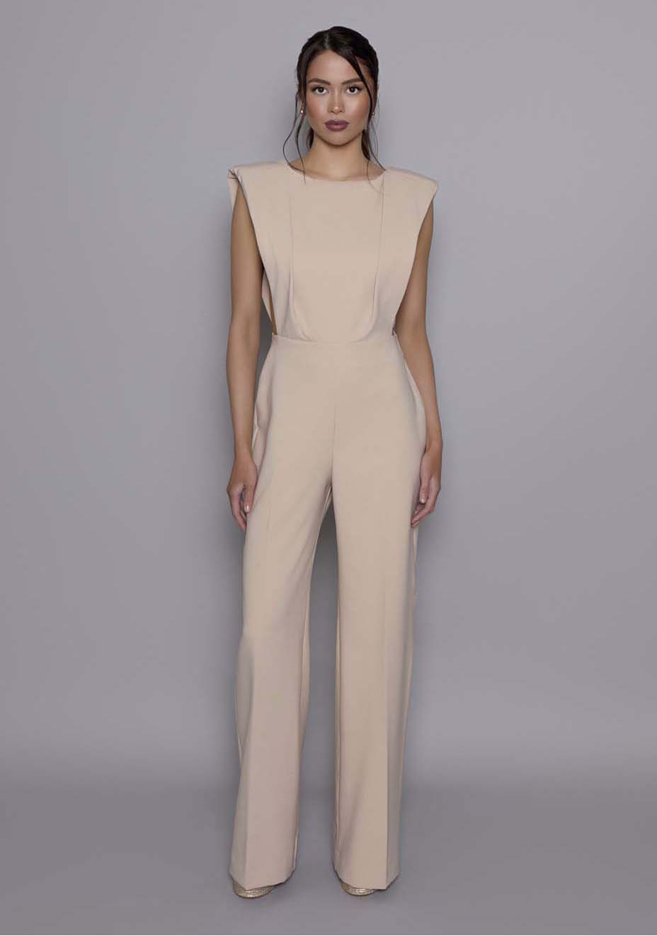 Model wearing the VSPR Bronte Stone open-back Jumpsuit featuring structured shoulders, a sculpted bodice, and wide-leg trousers, standing against a gray studio backdrop.