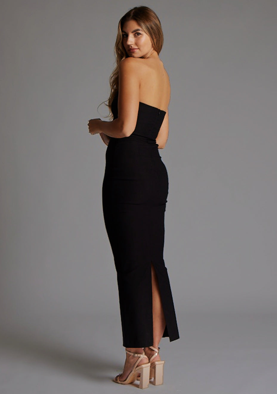 Side back image of a model styled in the Vesper Calix Bandeau Maxi Dress in Black, revealing the sleek strapless structure and elegant length.