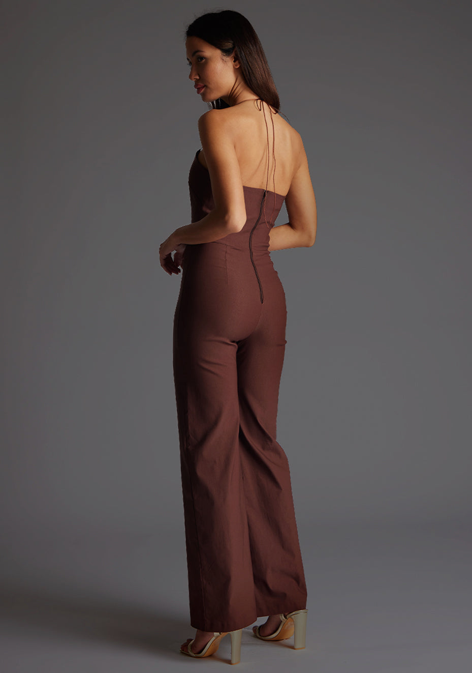 Side back image of a model wearing the Vesper Cassidy Halterneck Wide Leg Jumpsuit in Chocolate, showcasing the graceful halter tie detail at the back.