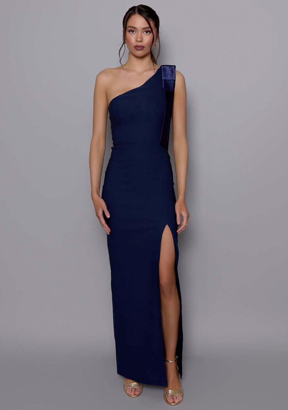 Model wearing the VSPR Cedar navy maxi dress with an asymmetric one-shoulder neckline and satin bow detail, featuring a slim fit and thigh-high split, against a grey studio backdrop.