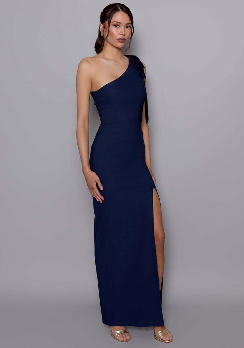 Side-front view of the VSPR Cedar navy maxi dress highlighting the draped one-shoulder silhouette, bow accent, and contouring fit through the waist and hips.