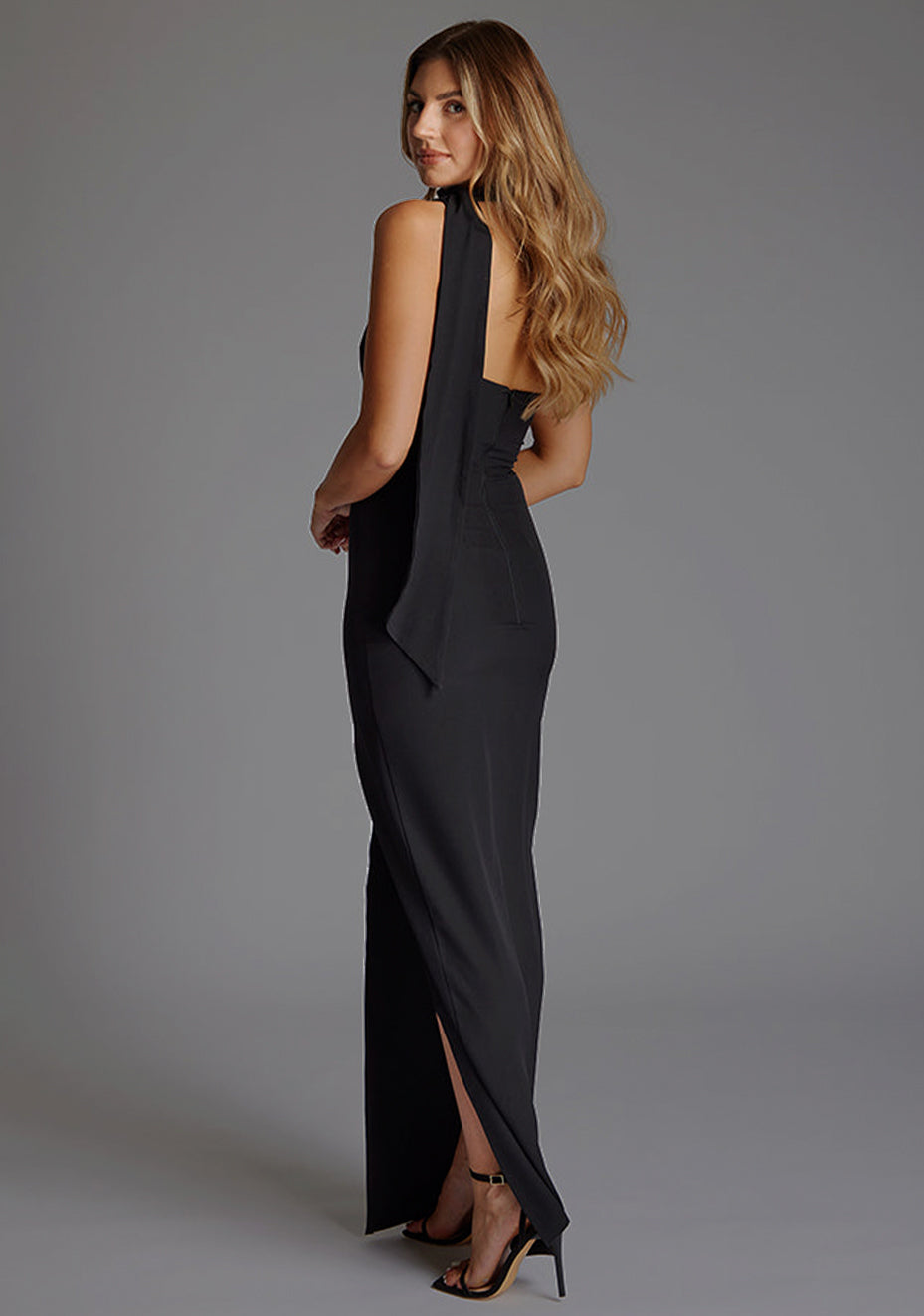 Side back image of a model wearing a black maxi evening dress with asymmetric neckline, front skirt split and statement collar drape detail. The design featured is the Vesper Cordula Scarf Detail One Shoulder Maxi Dress Black.