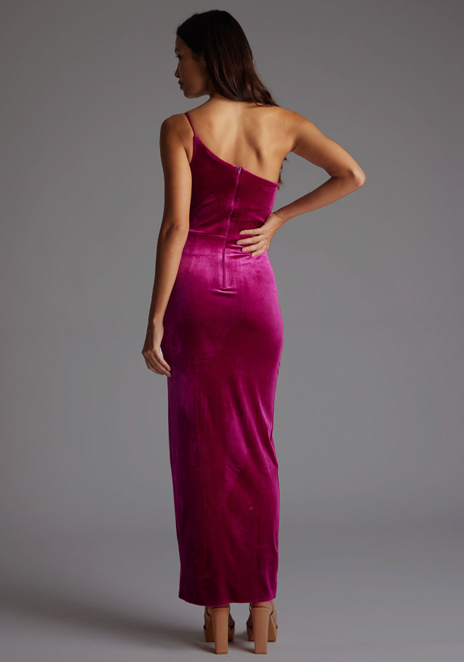 Back image model wearing a magenta maxi dress with a flattering asymmetric cut-out neckline, finished with a one shoulder strappy detail and front skirt split. The design featured is the Vesper Daniella One Shoulder Velvet Maxi Dress Magenta.