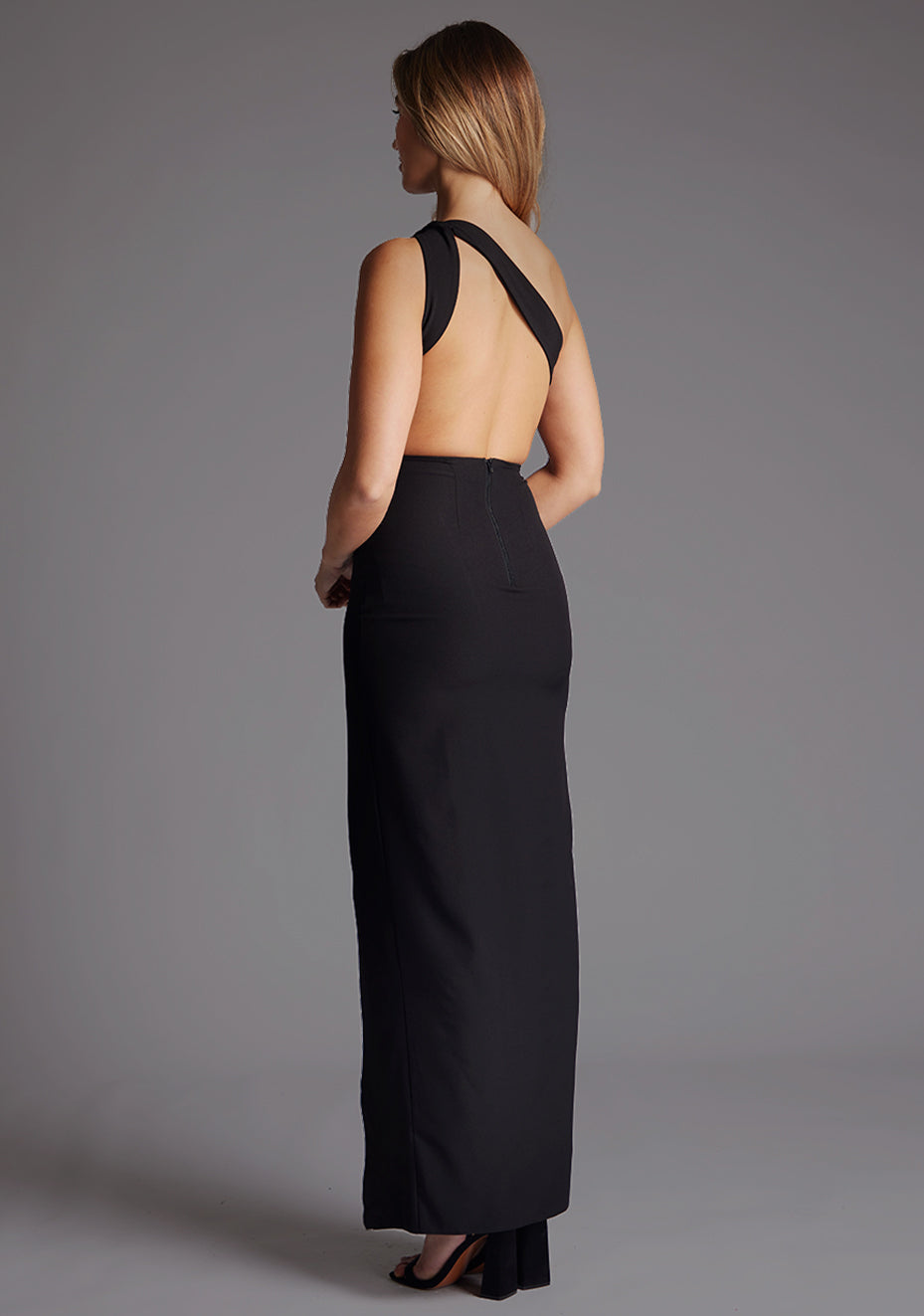 Side back image of a model wearing the Vesper Deva One Shoulder Open Back Maxi Dress in Black, revealing the contemporary open back design and flowing skirt.