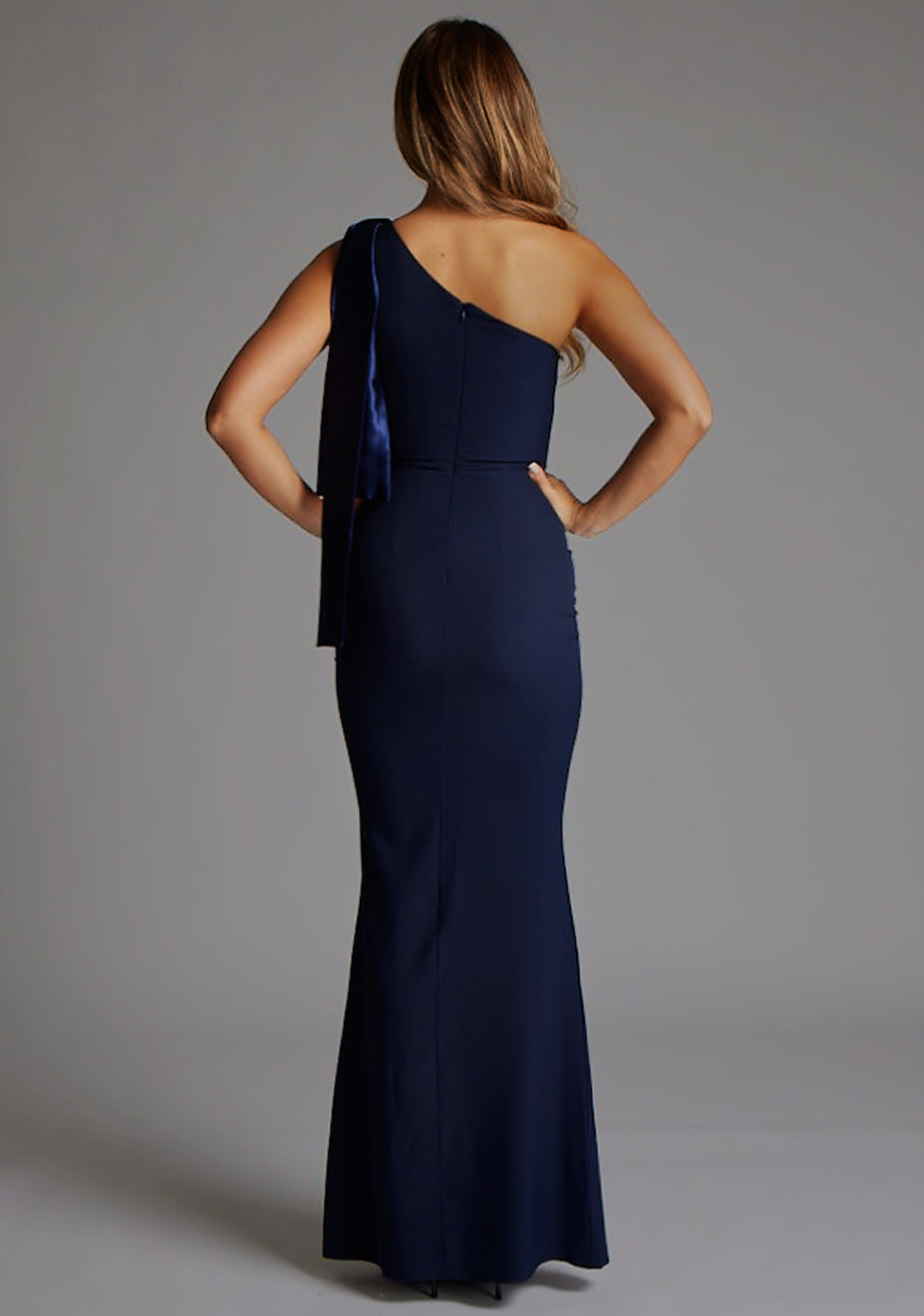 Back image of a blonde model wearing a Navy Maxi Dress with an asymmetric neckline and one-shoulder. The design features the Vesper Deven One Shoulder Fishtail Maxi Dress in Navy.