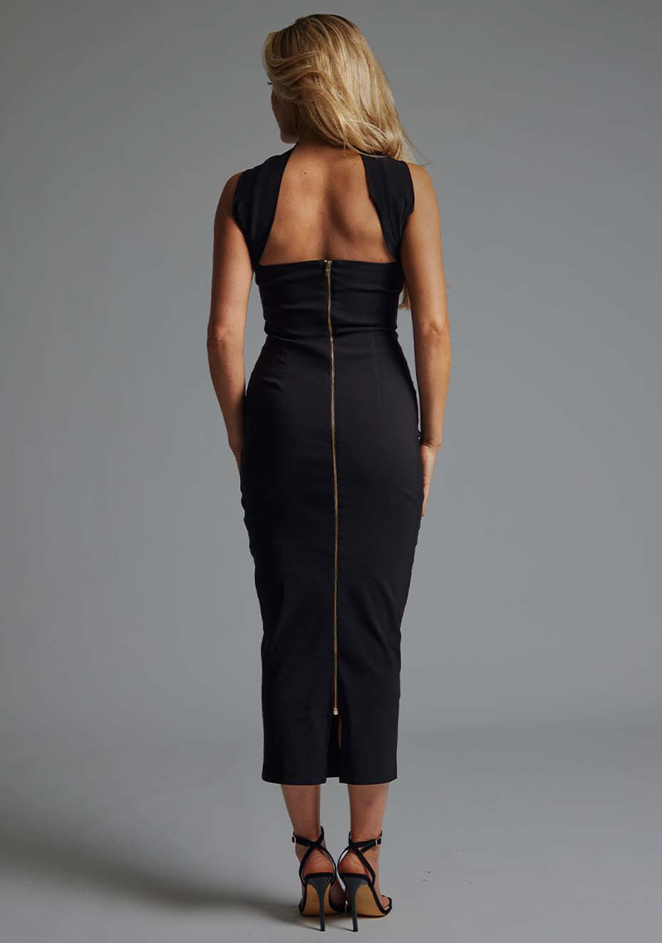 Back image of blonde model wearing a black collar detail midaxi dress with a straight neckline and collar detailing at the neck with straps at the back and statement gold centre back zip. The design featured Vesper Dion Collar Detail Midaxi Dress in Black.