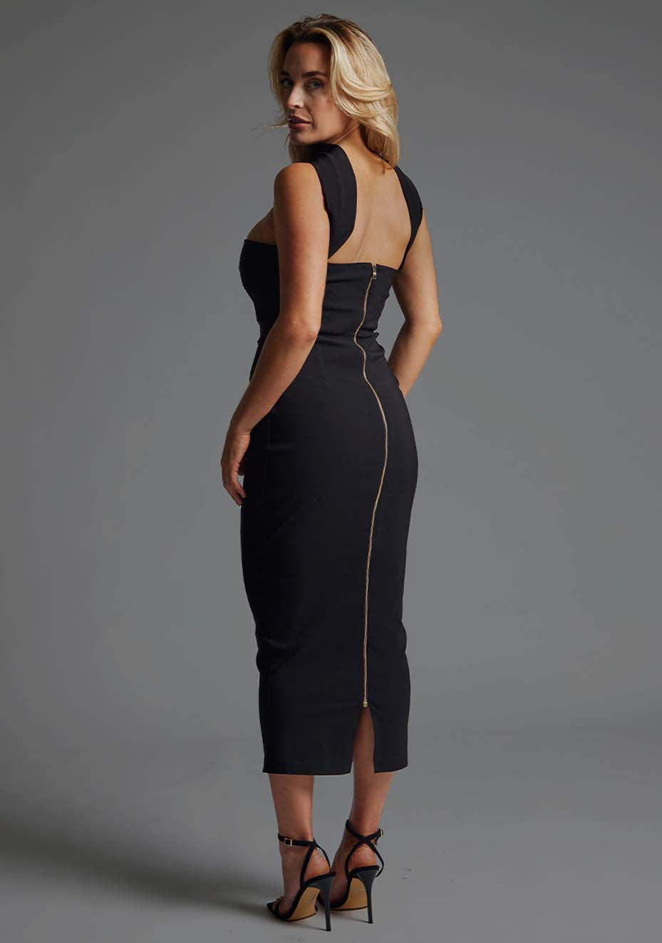 Side back image of blonde model wearing a black collar detail midaxi dress with a straight neckline and collar detailing at the neck with straps at the back and statement gold centre back zip. The design featured Vesper Dion Collar Detail Midaxi Dress in Black.