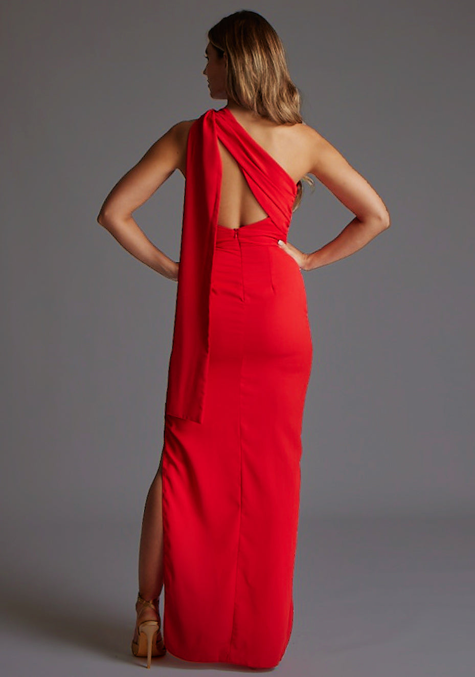 Back image of a model wearing a red maxi dress with a one shoulder design with draped fabric at shoulder and statement asymmetric back with cut-out details and front skirt split. The design featured is the Vesper Drew One Shoulder Drape Detail Maxi Dress in Red.