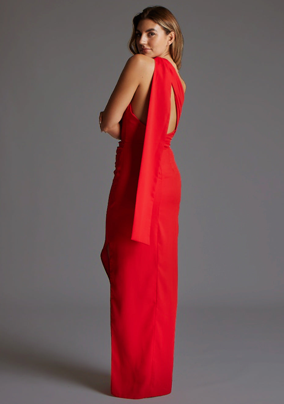 Side back image of a model wearing a red maxi dress with a one shoulder design with draped fabric at shoulder and statement asymmetric back with cut-out details and front skirt split. The design featured is the Vesper Drew One Shoulder Drape Detail Maxi Dress in Red.