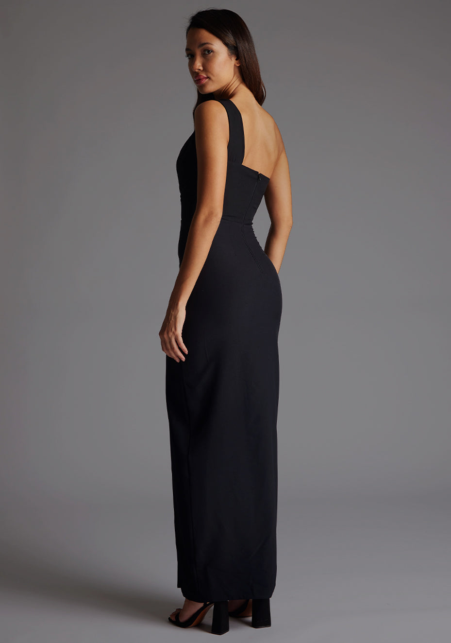 Side back image of the model wearing a black maxi dress with a sleek one-shoulder silhouette, contemporary front cut-out and front skirt split for effortless chic. The design features Vesper Ellis Cut-Out One-Shoulder Maxi Dress in Black.