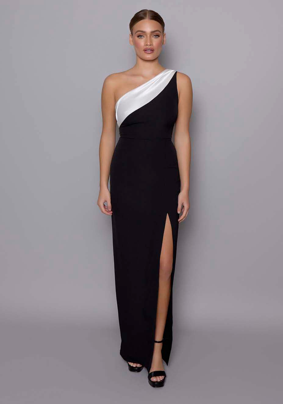 Model wearing the VSPR Fliss monochrome maxi dress with one‑shoulder neckline and ivory satin band, body‑con fit flowing to a thigh‑high split, posed against a neutral grey studio backdrop.