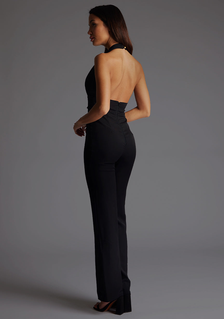 Side back image of the model wearing a black jumpsuit with a chic plunge neckline and its open back adds a hint of allure. The design featured is the Vesper Flynn Plunge Neckline Wide Leg Jumpsuit in Black.