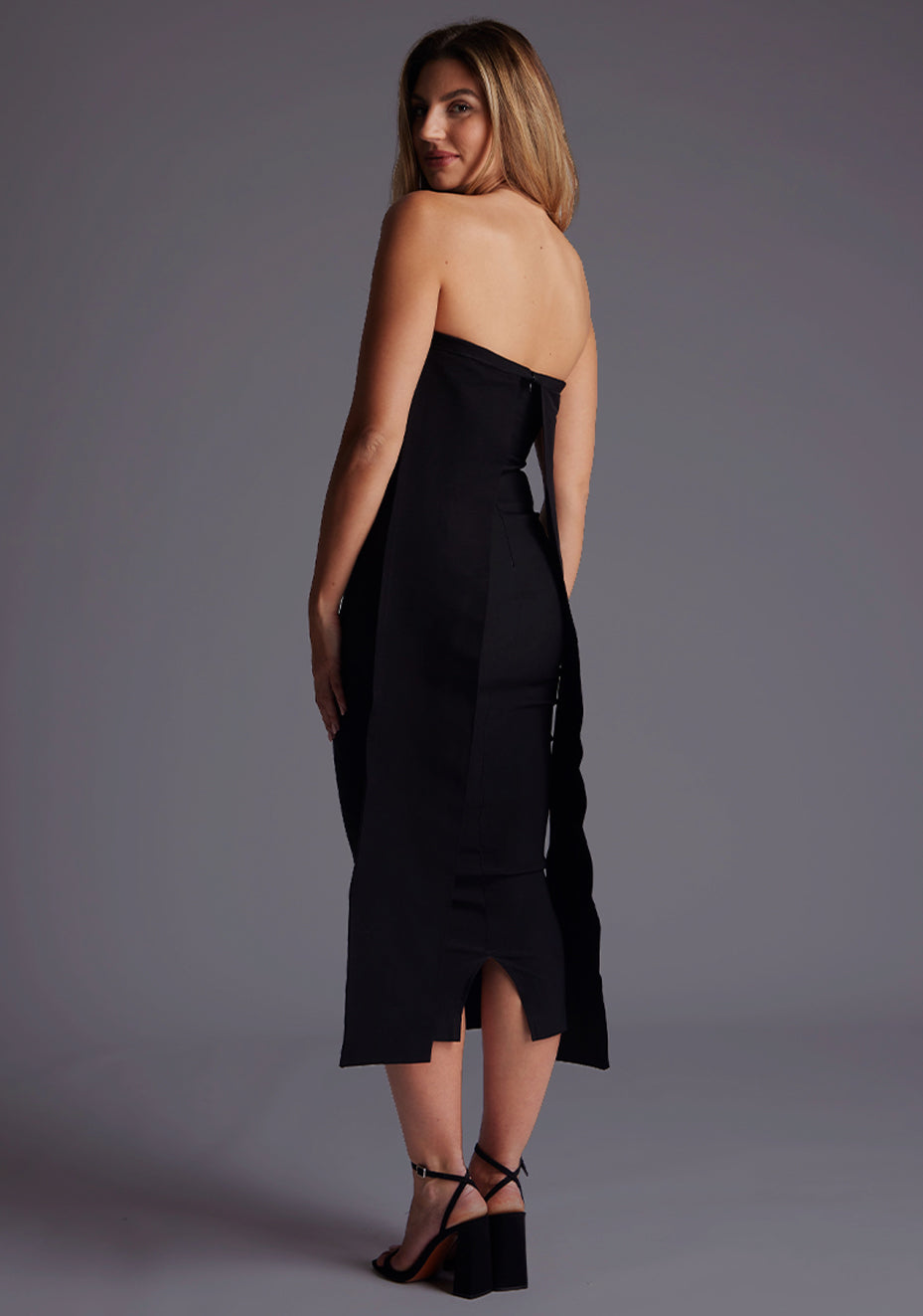 Side back image of a model wearing a black midaxi dress with a featuring a straight neckline, mid-length hem and flowing fabric at beck. The design featured is the Vesper Gabriel Black Midaxi Dress