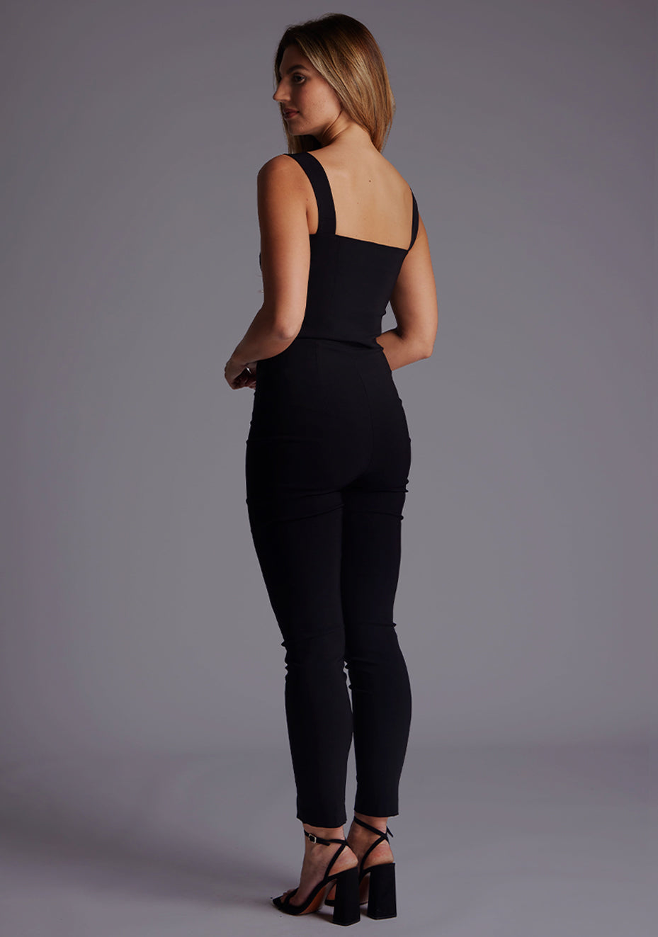 Side back image of the model wearing pine green top with a sweetheart neckline and wide straps create a flattering silhouette, while the circular cut outs at the hem add a touch of edge and statement front zip. The design featured is the Vesper Juno Sweetheart Crop Top in Black with Vesper Raven High Waist Trousers in Black.