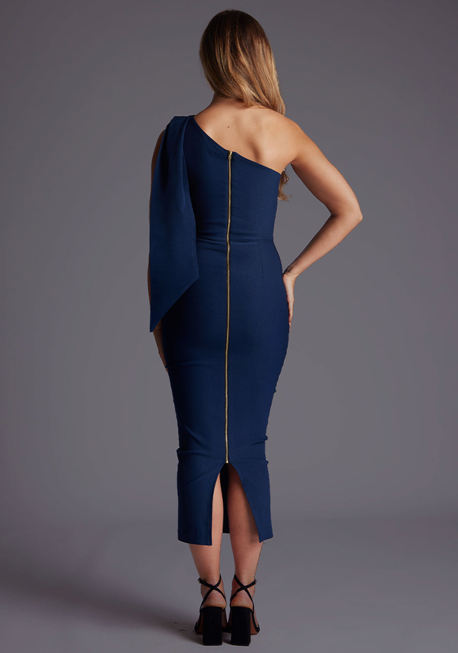 Back image of a model wearing a navy midaxi dress with a chic one-shoulder design and an asymmetric silhouette, it effortlessly captivates with its modern flair. The design featured is the Vesper Katrina One Shoulder Drape Midaxi Dress in Navy.
