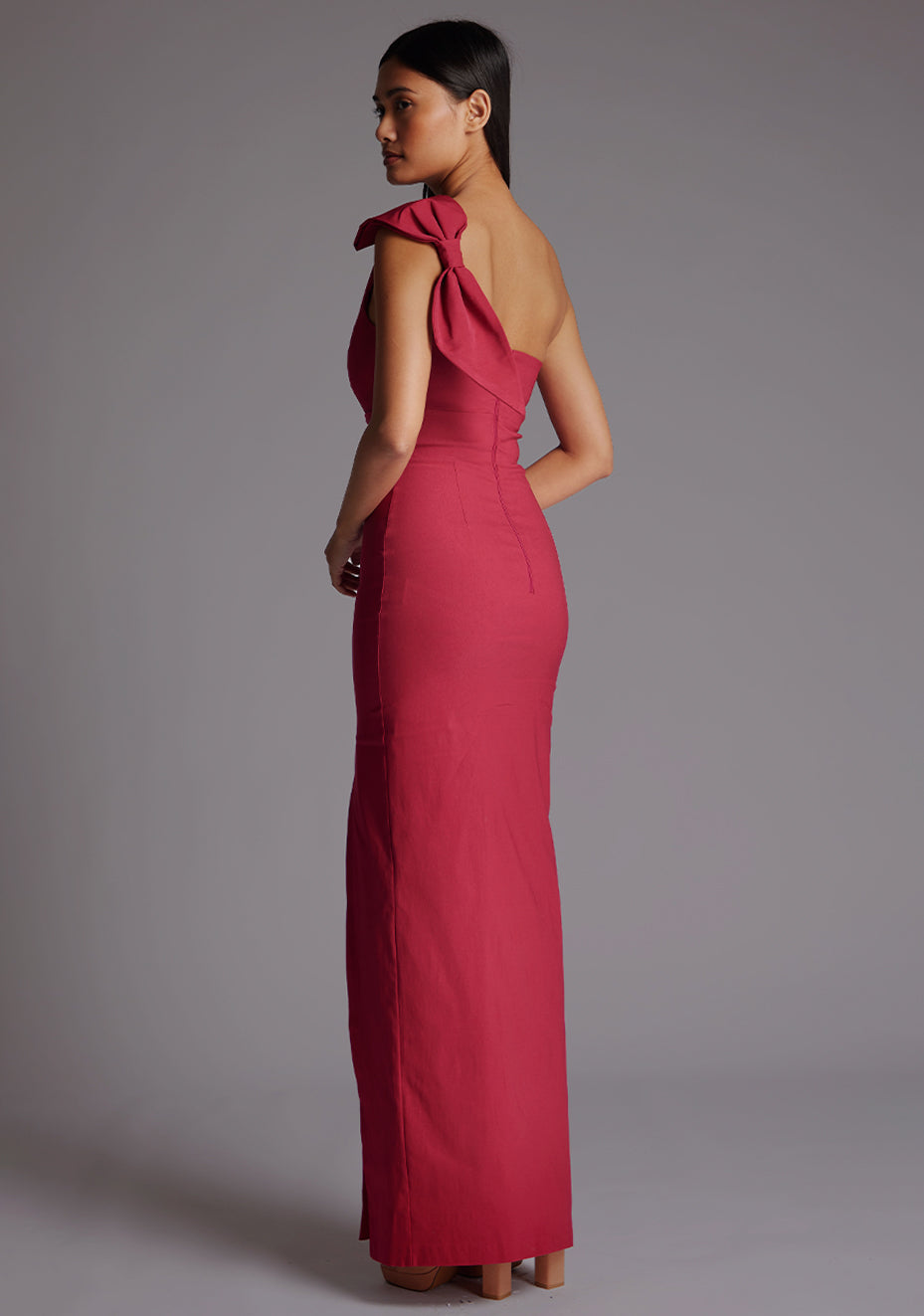Side back image of a model wearing a raspberry maxi dress, featuring an asymmetric neckline, one shoulder with statement bow detail, and a front skirt split. The dress feature is the Vesper Keegan One Shoulder Maxi Dress in Raspberry.
