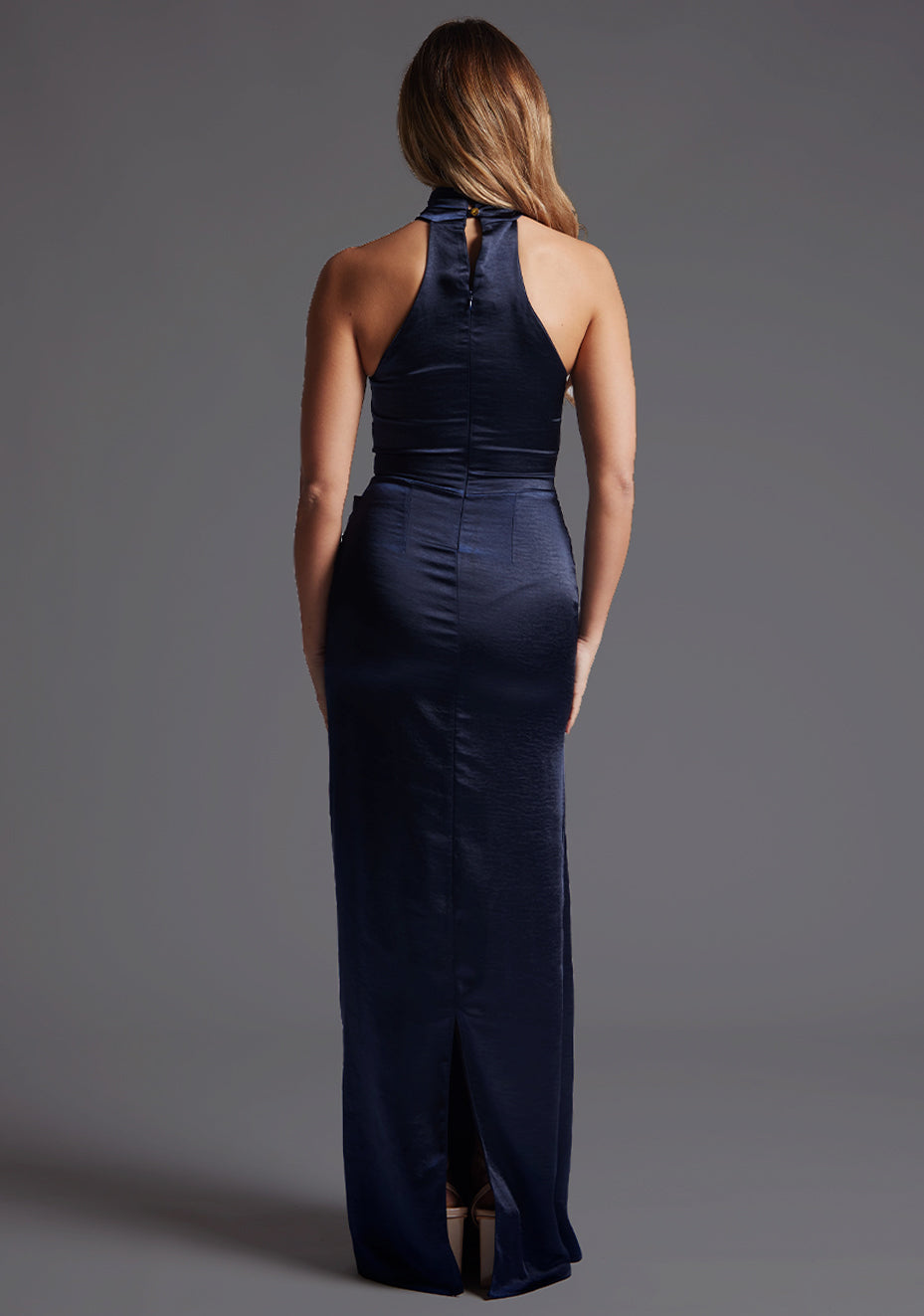 Back image of model wearing a lake navy maxi dress with a flattering halter neckline, crafted in a rich navy hue, it exudes timeless charm, back slit and invisible centre back zip. The design featured is Vesper Lake Halter Neckline Maxi Dress in Navy.