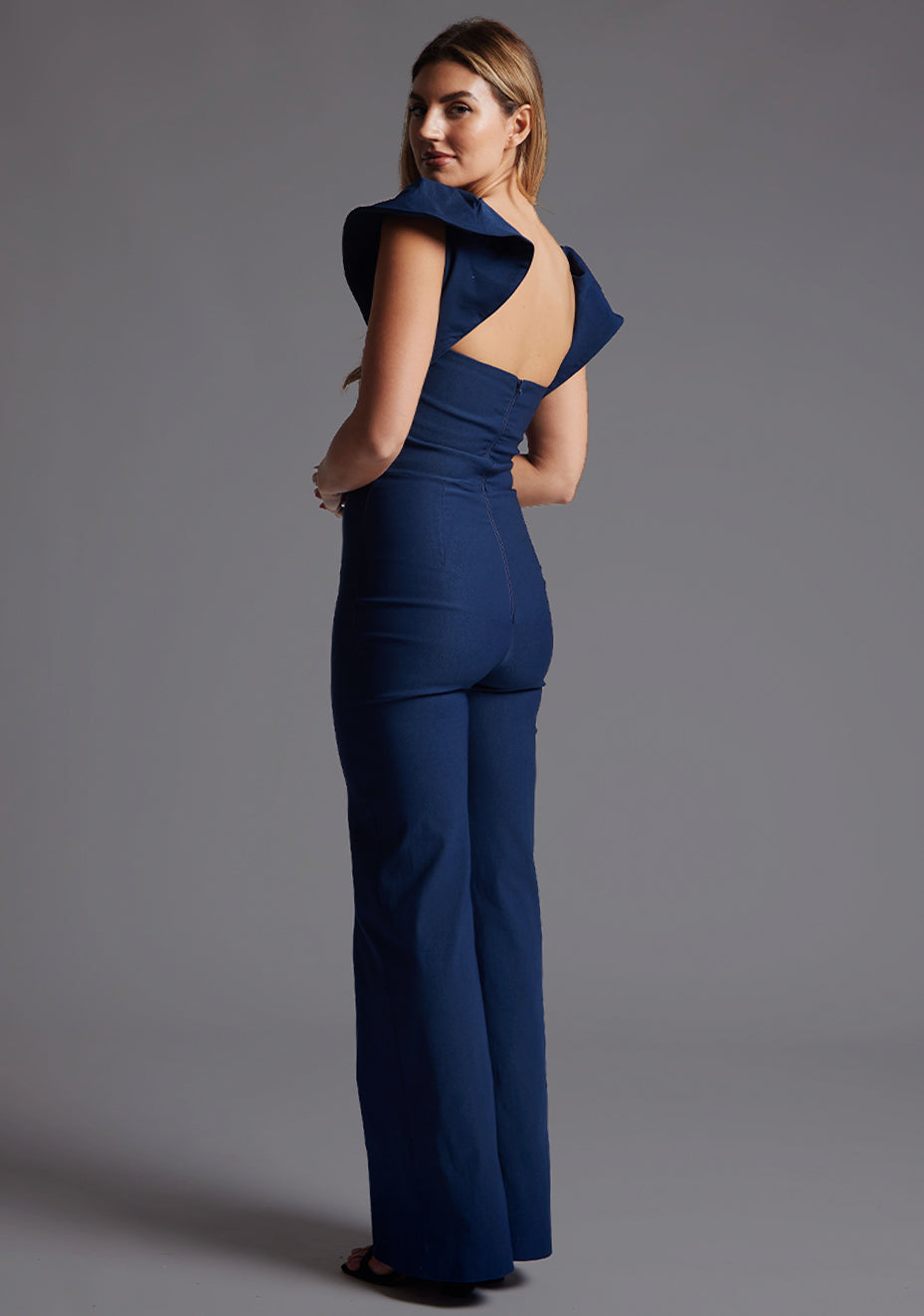 Side back image of a model wearing the Vesper Leigh Wide Leg Frill Sleeve Jumpsuit in Navy, showing the smooth back fit and flowing wide-leg shape.