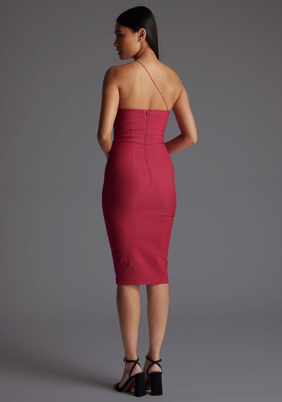 Back quarter image of the model wearing an raspberry one shoulder midi dress with a asymmetrical neckline with one strap, bodycon silhouette and discreet back zip for seamless fit. The design featured is the Vesper Leslie One Shoulder Midi Dress in Raspberry.