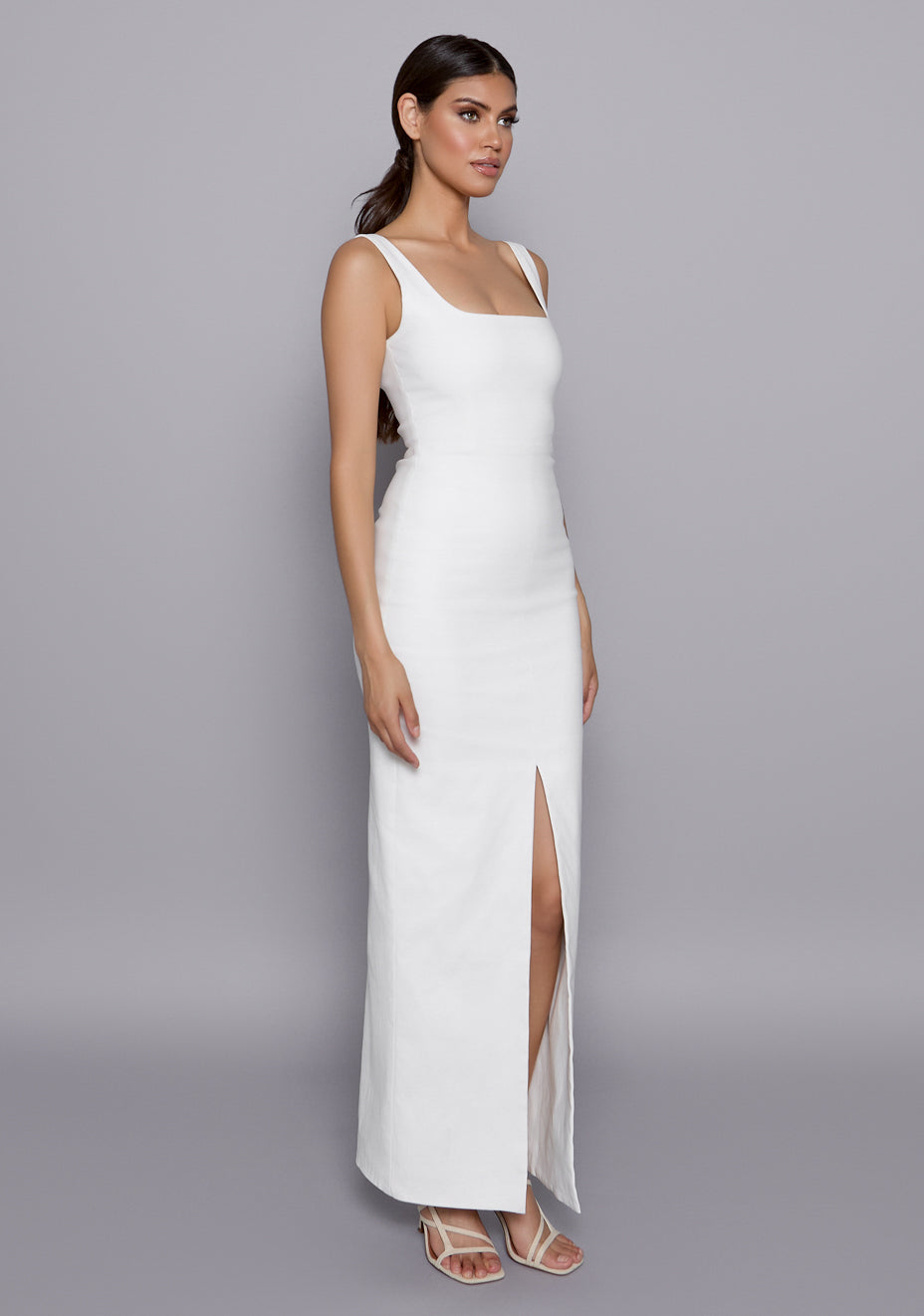 Side-front view of the VSPR Ria Ivory Maxi Dress revealing the body-hugging fit, front slit opening, and contoured silhouette accentuated by wide straps.