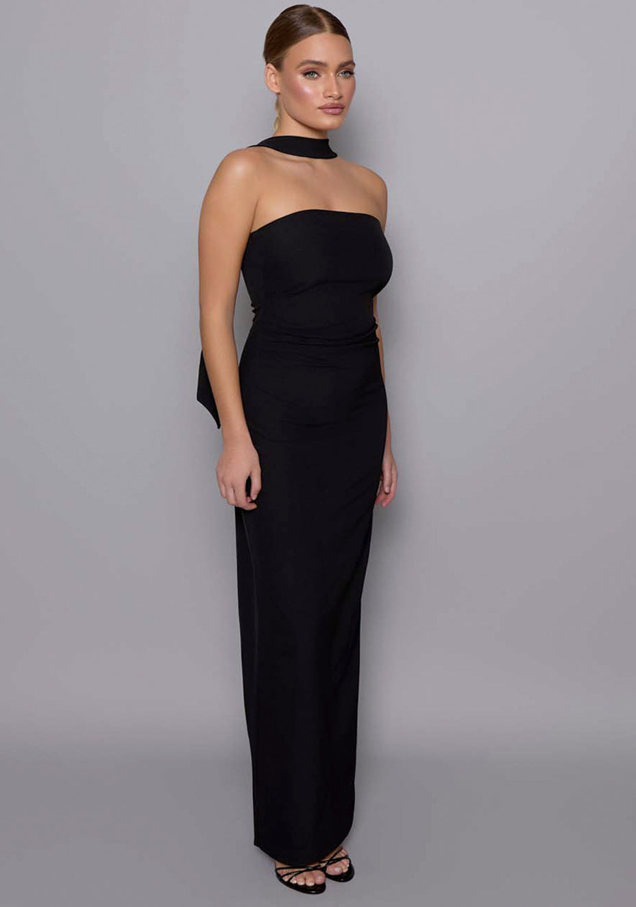 Side-front view of the VSPR Tiffany black maxi dress showcasing the curve-enhancing ruched waist, bandeau neckline, and scarf halter draping over the shoulder.