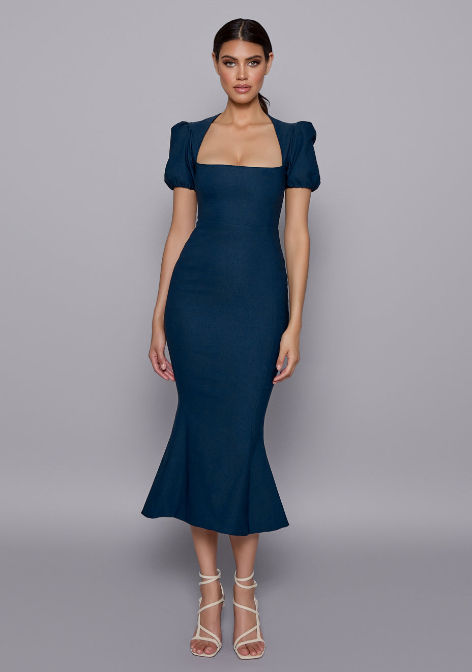 Model wearing the VSPR Valencia Fishtail Navy Midaxi Dress with a square neckline, puffed short sleeves, and fishtail hem, standing against a neutral studio backdrop.