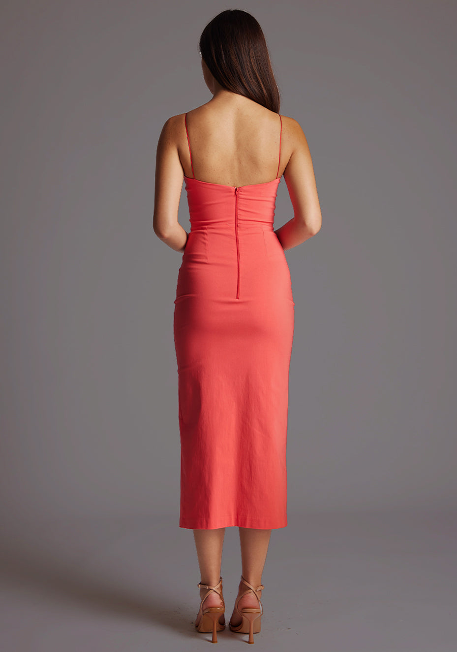 Back image of a model wearing a coral midaxi dress with slender straps, a graceful V-neckline, and a captivating front skirt split. The design featured is the Vesper Bronwen Coral Midaxi Dress.