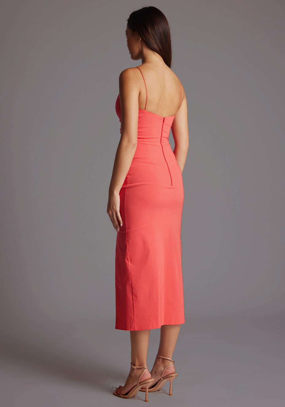 Back quarter image of a model wearing a coral midaxi dress with slender straps, a graceful V-neckline, and a captivating front skirt split. The design featured is the Vesper Bronwen Coral Midaxi Dress.