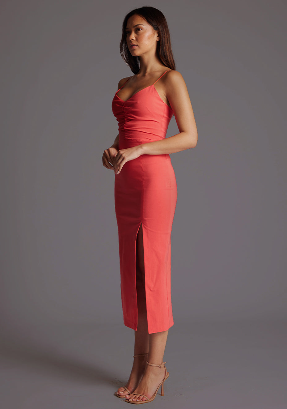 Front quarter image of a model wearing a coral midaxi dress with slender straps, a graceful V-neckline, and a captivating front skirt split. The design featured is the Vesper Bronwen Coral Midaxi Dress.