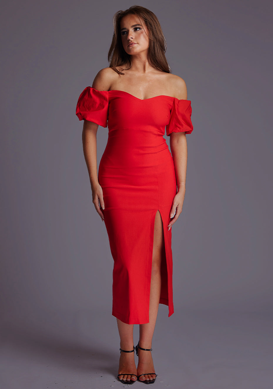 Front image of a model wearing a red midaxi dress with a off shoulder design with sweetheart neckline, puffy bands at the top of arms and front skirt split. The design featured is the Vesper Lottie Red Midaxi Dress