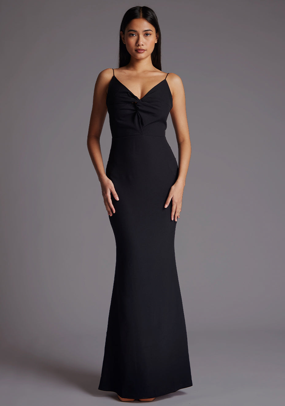 Front image of a model wearing a black maxi dress with a v-neckline, thin straps with twist front detail at bust, low back and subtle flair at hem. The design featured is the Vesper Marlowe Black Maxi Dress