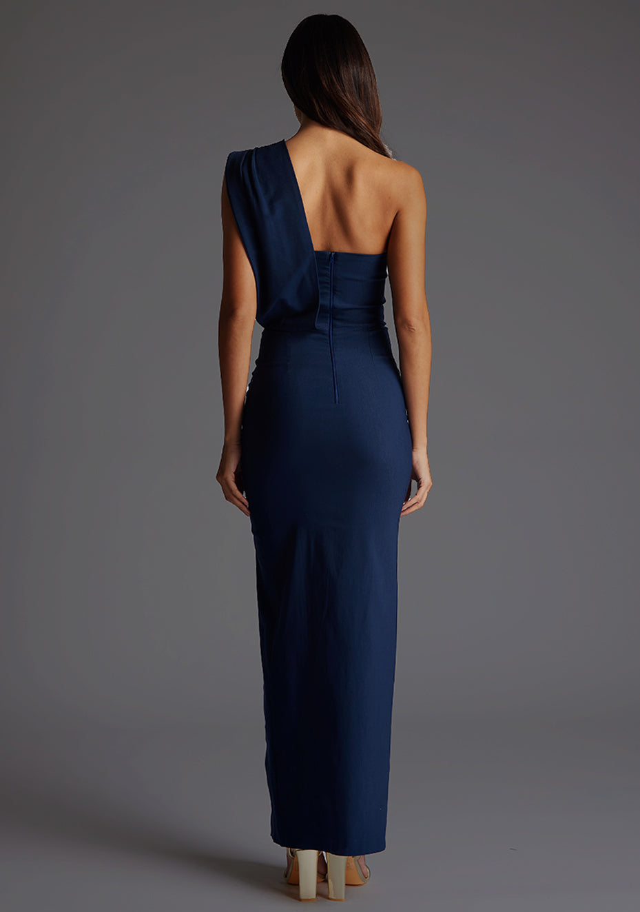 Back image of a model wearing a navy one shoulder maxi dress with a straight neckline with an asymmetric one shoulder draped fabric with pleat detailing and front skirt split. The design featured is the Vesper Marni Navy One Shoulder Maxi Dress