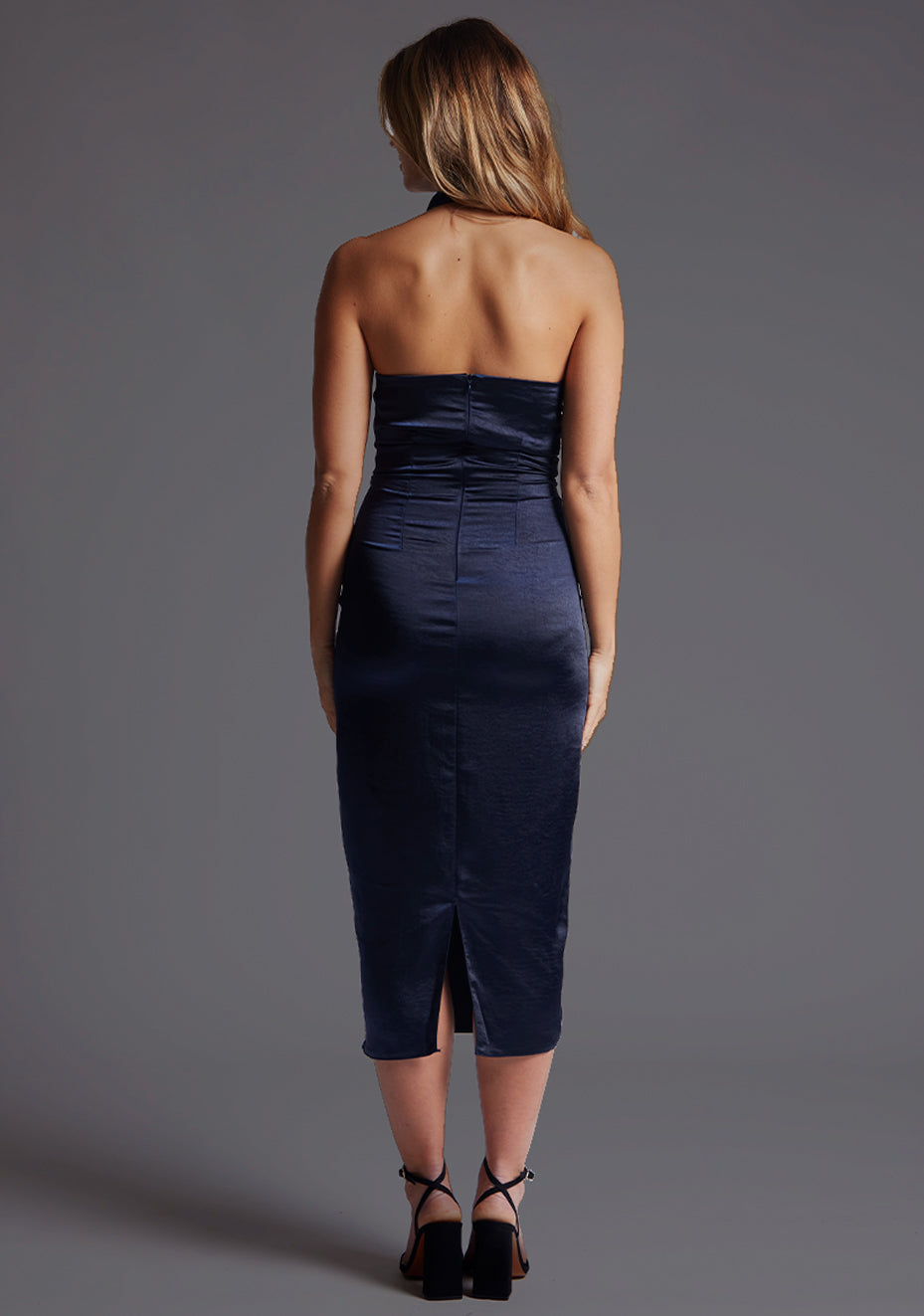 Back image of a blonde model wearing a navy midaxi dress with a asymmetric neckline with one-shoulder design, captivating cowl design and slim fit detail. The design featured is the Vesper Mason Navy Midaxi Dress.