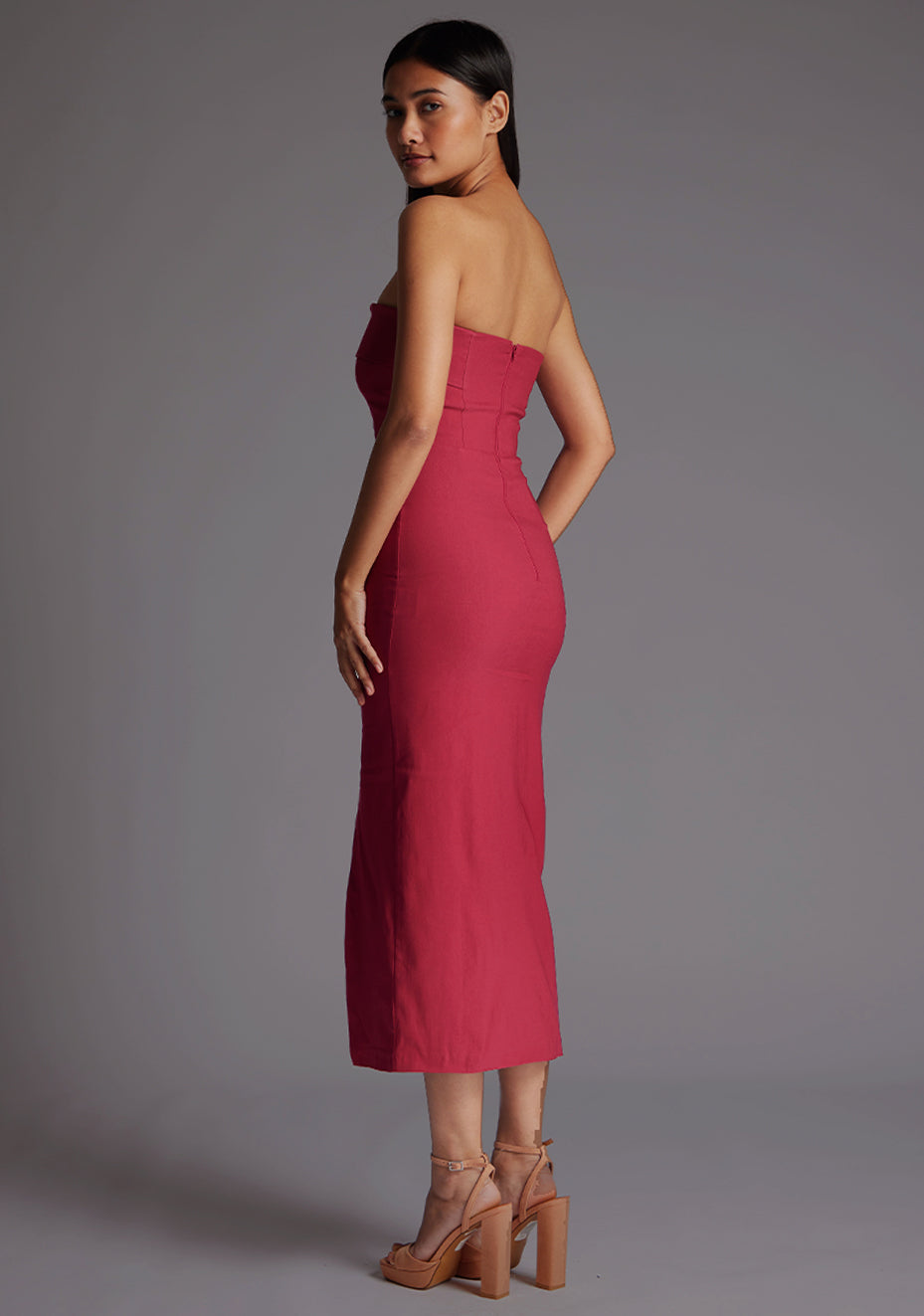 Side back image of a model wearing a raspberry midaxi dress with a straight-across strapless design, side slit and bodycon fit. The design featured is the Vesper Monica Strapless Midaxi Dress in Raspberry.