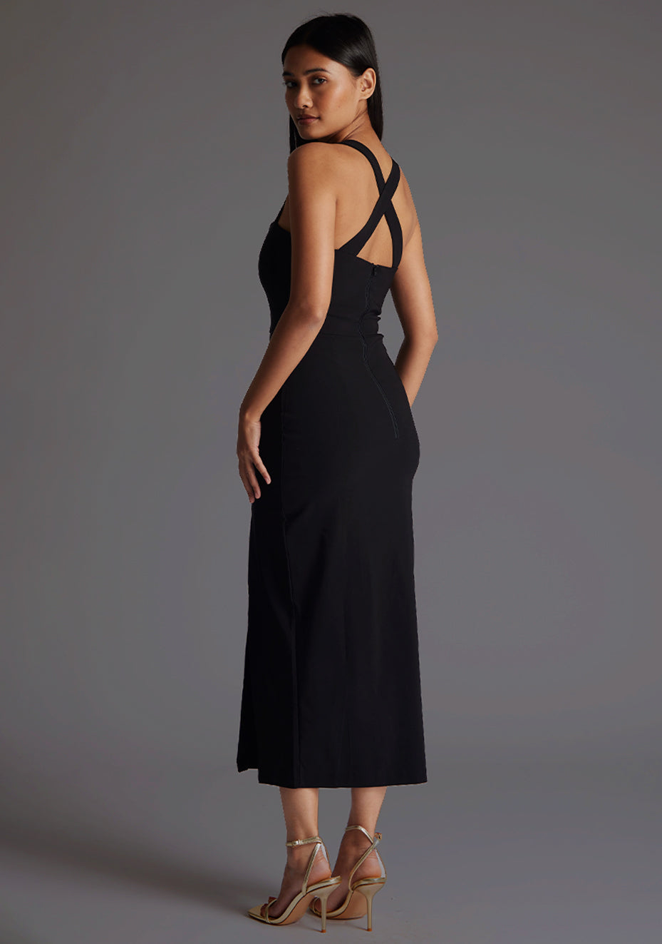 Back quarter image of model wearing a black sweetheart midaxi dress with a plunge sweetheart neckline with wide straps that cross at the back and a front skirt split. The design featured is the Vesper Poppy Black Sweetheart Midaxi Dress
