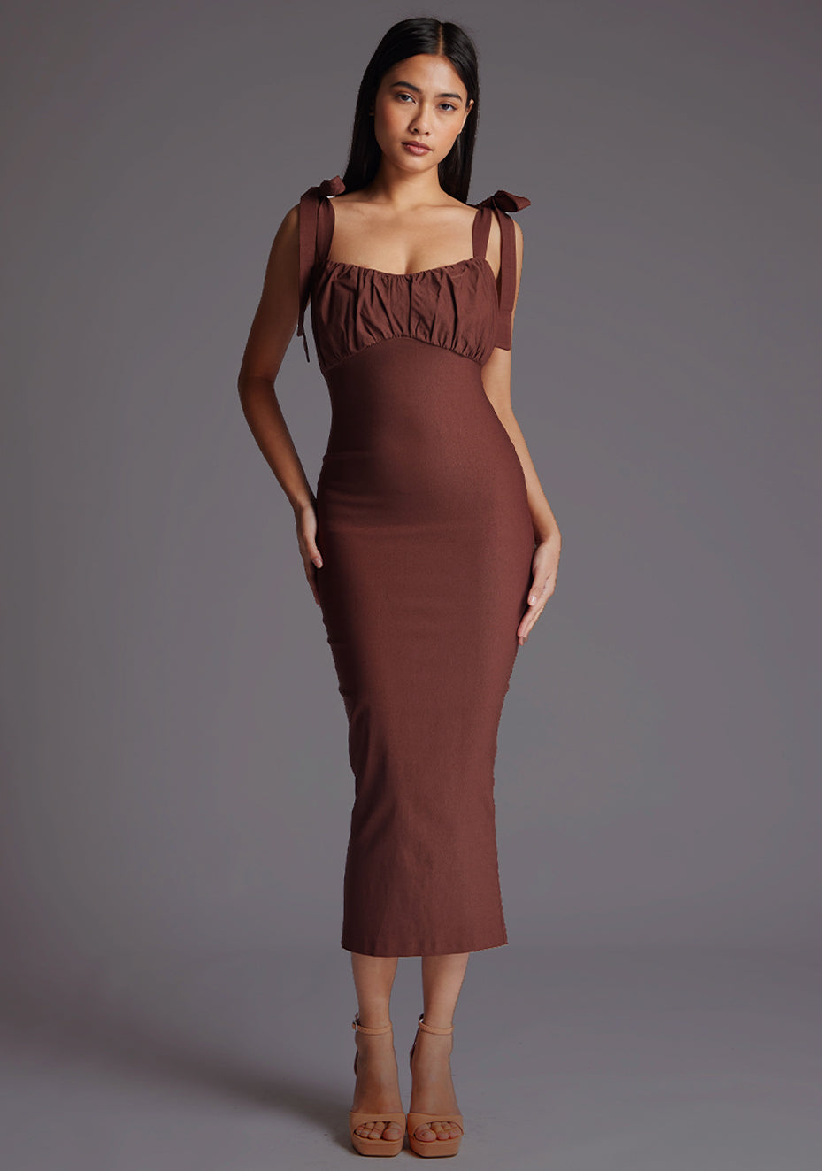 Front image of a model wearing a chocolate midaxi dress with a subtle sweetheart neckline, self-tie bow straps, pleating detail at bust, bodycon fit with statement gold zip at centre back .The design featured is the Vesper Rapunzel Chocolate Midaxi Dress.
