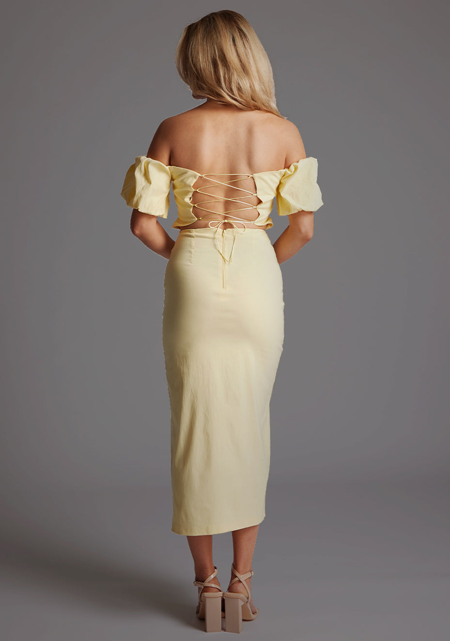 Back image of a blonde model wearing a yellow bardot midaxi dress with a subtle sweetheart neckline, statement puff sleeves, a front skirt split and lace-up detail at the back. The design featured is the Vesper River Yellow Bardot Midaxi Dress